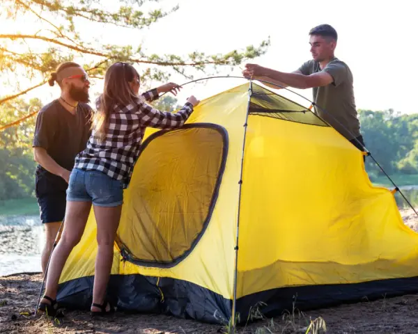 Friends setting up a bright yellow tent by a lake, learning how to choose a campsite with a beautiful sunset.