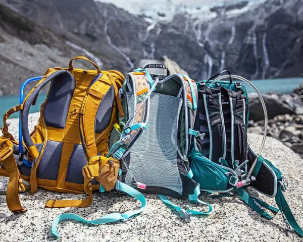 Three Osprey backpacks sit on rocks by a lake, ready for a hike. Learn how to pack a day pack for hiking with these essential tips.