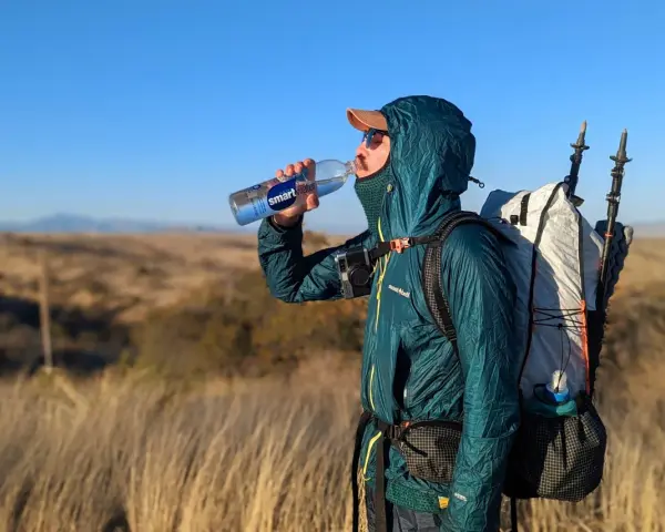 A hiker takes a refreshing drink from a Smartwater bottle while carrying a large backpack and trekking poles.