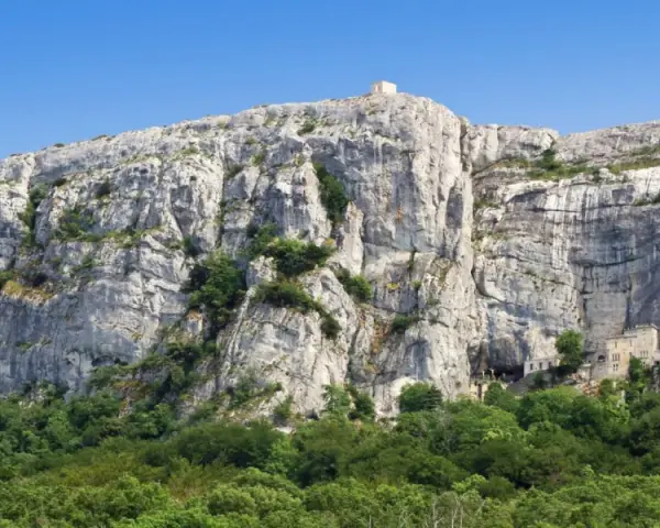 A majestic cliff face rises above a lush green forest, with a small building nestled into the rock at Sainte Baume.