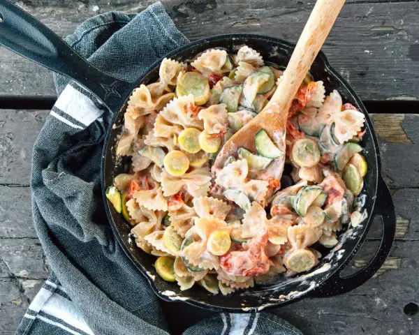 A hearty camping pasta dish with farfalle, zucchini, and tomatoes in a creamy sauce, served in a cast-iron skillet.