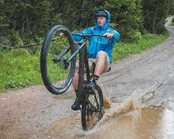 A mountain biker in a blue jacket performs a wheelie through a large puddle, splashing water as it rains heavily on a forest trail.