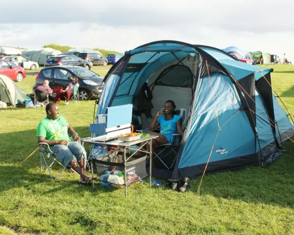 A couple enjoys a meal at their campsite, a perfect example of how to camp. They sit in chairs beside a blue tent, with food laid out on a portable table.