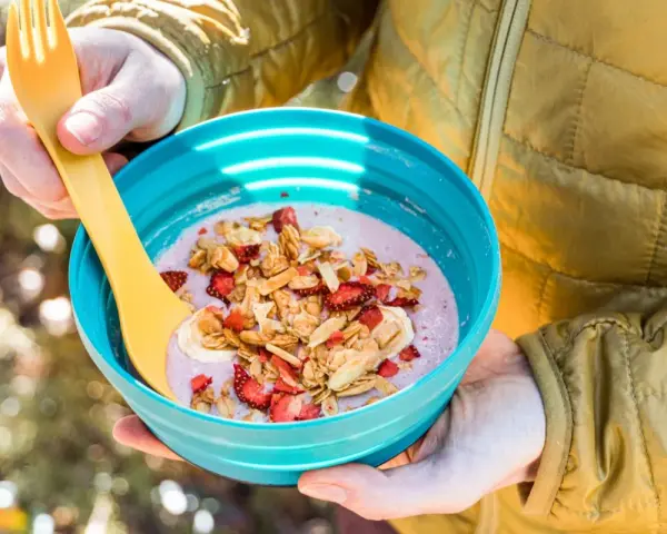 A person in a yellow jacket holds a blue bowl filled with a pink smoothie topped with granola, dried strawberries, and banana slices. Perfect for camping breakfast ideas.