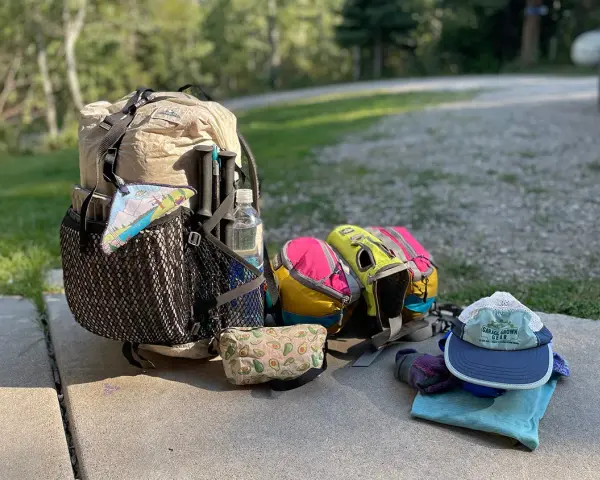 Ready for adventure! A large backpack, hiking poles, water bottle, and colorful pouches are laid out with camping gear, including a hat and gloves.