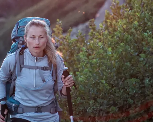 A woman with blonde hair, wearing a grey jacket and a large backpack, is backpacking hiking up a rocky, vegetated slope. She holds a trekking pole.