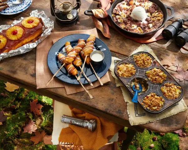 A spread of cozy camping food: pineapple upside-down cake, pigs in a blanket, apple crumble muffins, and a skillet brownie with ice cream.