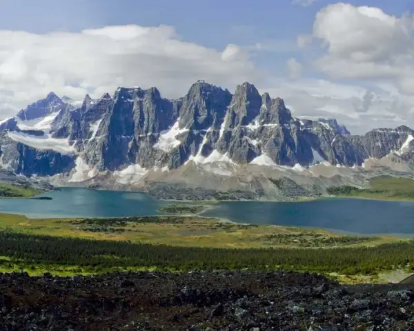 A vast mountain range with snow-capped peaks overlooks a serene blue lake. The Tonquin Valley Trail offers this breathtaking view of rugged beauty.