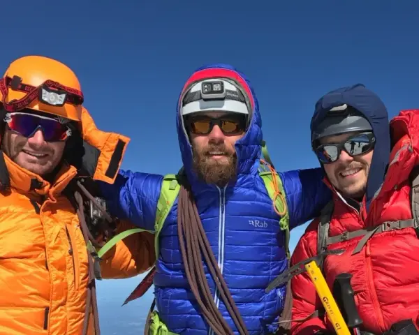 Three smiling mountaineers in bright down fill jackets and helmets pose on a sunny summit, ready for adventure.