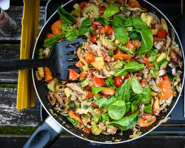 A colorful stir-fry with chicken, carrots, zucchini, and peppers, perfect for camp stove recipes. A black spatula is mixing the ingredients in a pan.