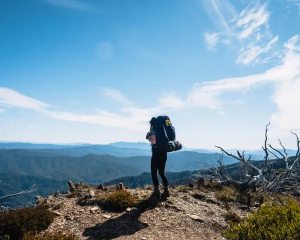 A lone hiker stands on a mountain ridge, ready for backpacking and hiking, with a vast expanse of blue mountains stretching to the horizon under a clear sky.