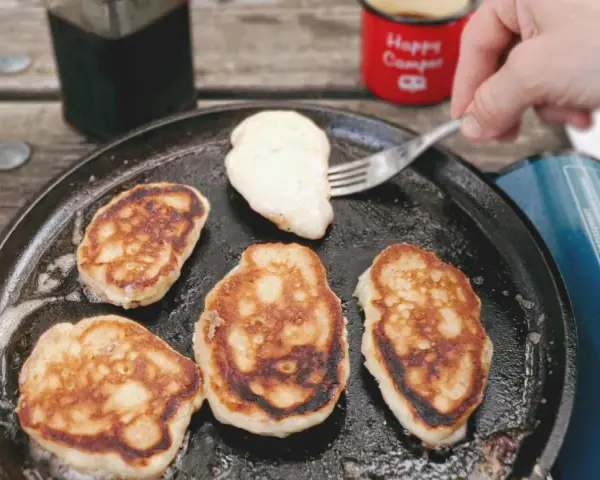 Frying pancakes in a cast iron skillet is a delicious answer to what to eat when camping. A fork lifts a fresh pancake, ready for syrup.