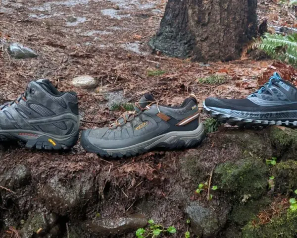 Three pairs of shoes on a wet forest floor: hiking boots, trail running shoes, and another pair of trail running shoes.