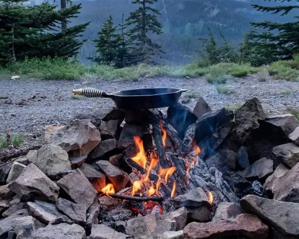 A cast iron skillet sits over a crackling campfire, ready for a delicious cast iron camping meal amidst a scenic mountain backdrop.