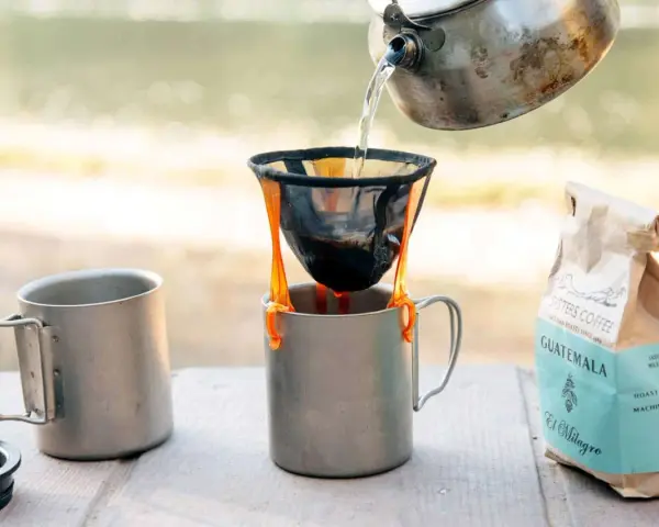 Making coffee while camping: hot water pours from a kettle into a pour-over filter, brewing coffee into a metal mug. A bag of Sisters Coffee Guatemala sits nearby.