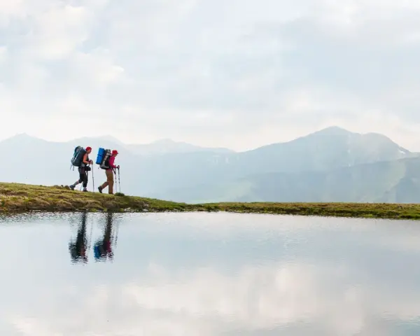 Two hikers with backpacks walk along a grassy ridge beside a calm lake, with misty mountains in the background. This image inspires thoughts on how to go backpacking.