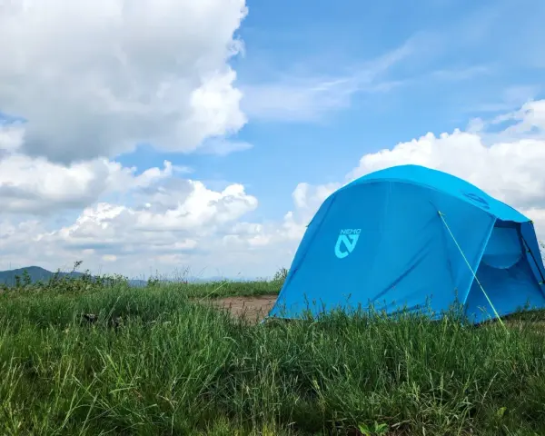 A bright blue Nemo tent stands in a grassy field under a cloudy sky. This Nemo Aurora tent is ready for adventure.