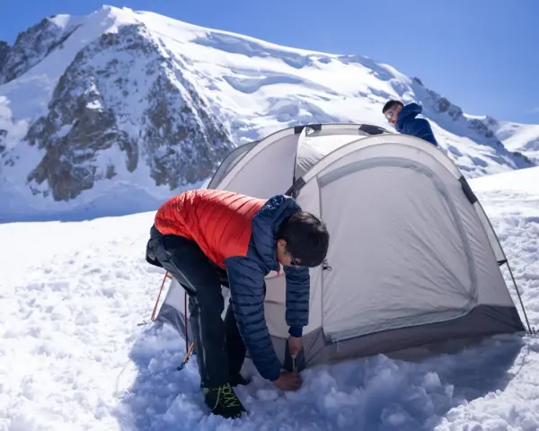 Two people set up a tent for winter camping amidst a snowy mountain landscape under a clear blue sky.