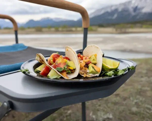 Delicious camping tacos with avocado, tomatoes, and lime, served on a plate on a portable table with mountains in the background.