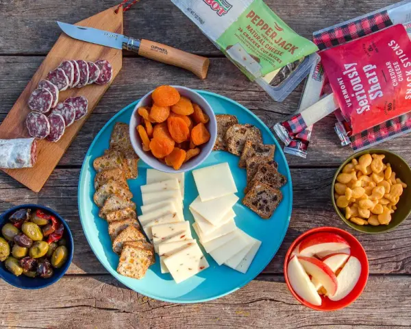 A delicious spread of camping snacks: sliced salami, cheese, crackers, dried apricots, olives, apple slices, and goldfish crackers.