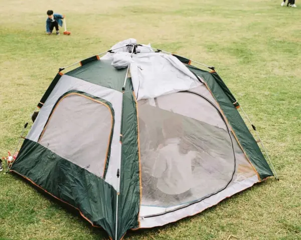 A green and gray tent is partially set up on a grassy field. A child plays in the background, learning how to set up a tent.