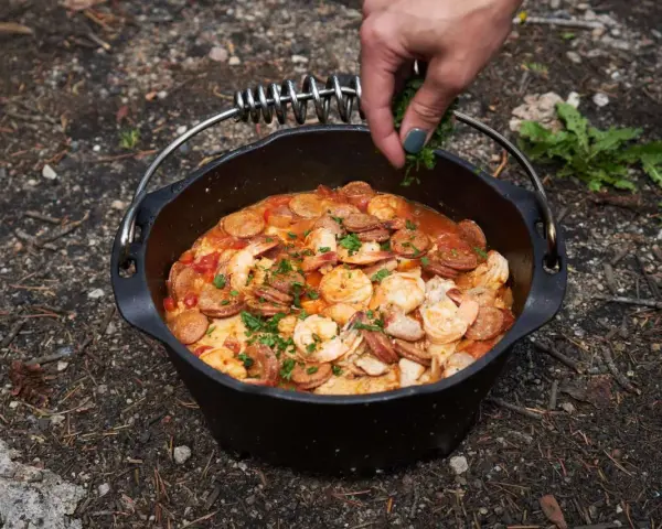 A hand garnishes a hearty dutch oven paella with fresh parsley. This campfire meal features shrimp, sausage, and rice in a rich tomato sauce.