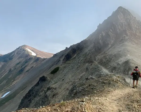 A hiker with a backpack and trekking poles walks along a narrow mountain ridge, a scenic path to learn how to get better at hiking.