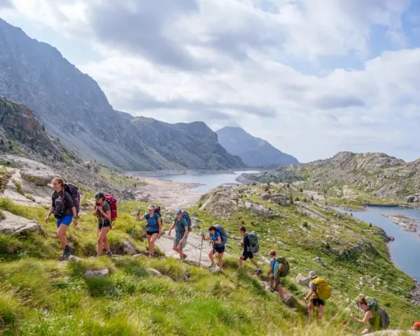 Hikers trek through a scenic mountain landscape, enjoying hiking the Pyrenees Spain. A chain of lakes and rugged peaks stretch into the distance under a cloudy sky.
