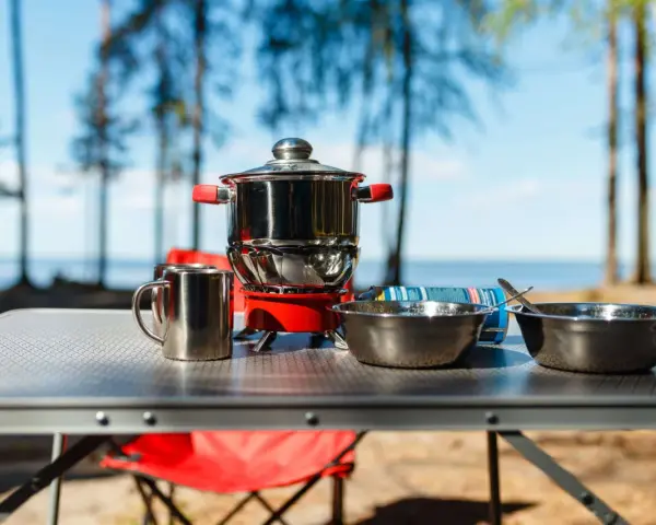 A complete camping kitchen setup with a stove, pots, bowls, and mugs on a table, ready for outdoor cooking by a lake.