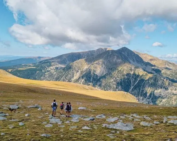 Hikers with backpacks traverse a rocky, grassy meadow on a pyrenees hut to hut hiking adventure, with majestic mountains under a cloudy sky.