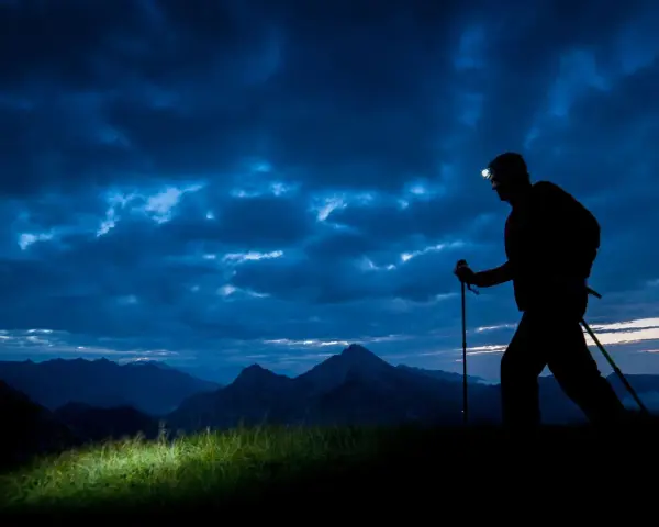 Silhouette of a hiker with a headlamp on, trekking poles in hand, on a night hike through a dramatic, cloudy sky over mountains.