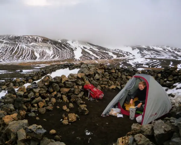 A hiker prepares a meal inside a tent at one of the Laugavegur trail huts, surrounded by snow-dusted mountains and a stone wall.