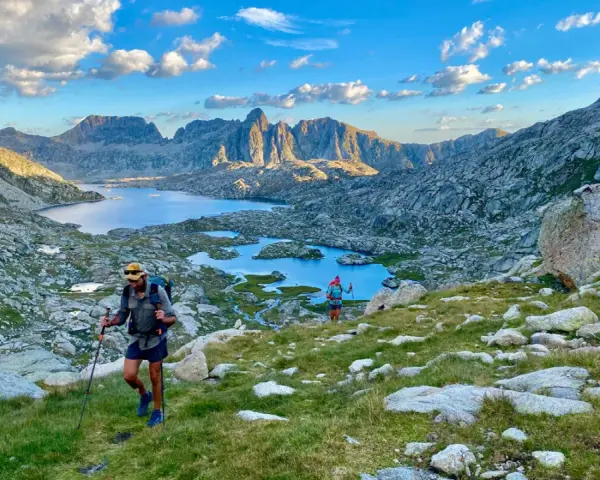 Hikers ascend a grassy slope on the HRP trail, with a stunning mountain lake and jagged peaks in the background under a cloudy sky.