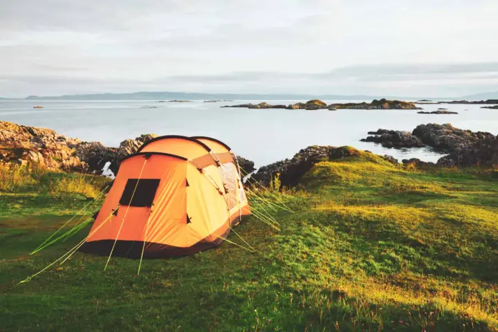 An orange tent is set up on a grassy hill overlooking a calm sea with rocky islands. This is a perfect spot for tent camp sites.