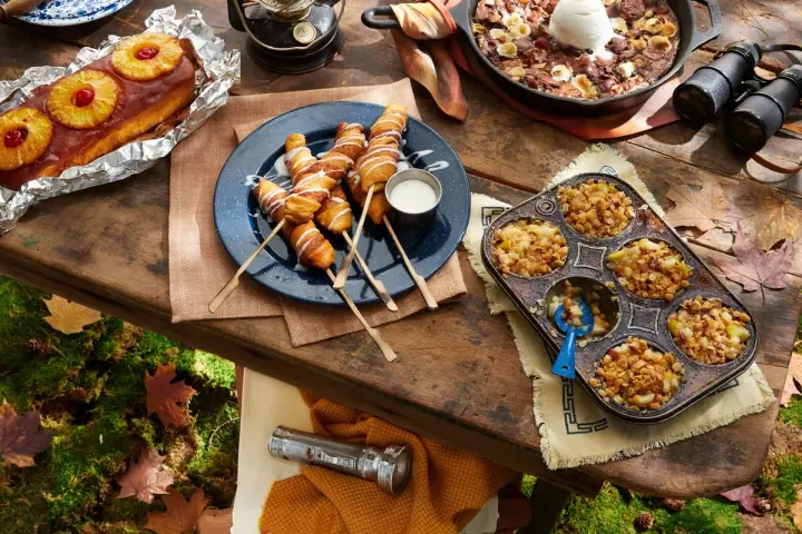 A spread of cozy camping food: pineapple upside-down cake, pigs in a blanket, apple crumble muffins, and a skillet brownie with ice cream.