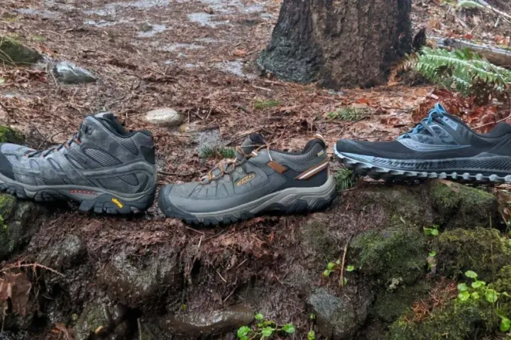 Three pairs of shoes on a wet forest floor: hiking boots, trail running shoes, and another pair of trail running shoes.