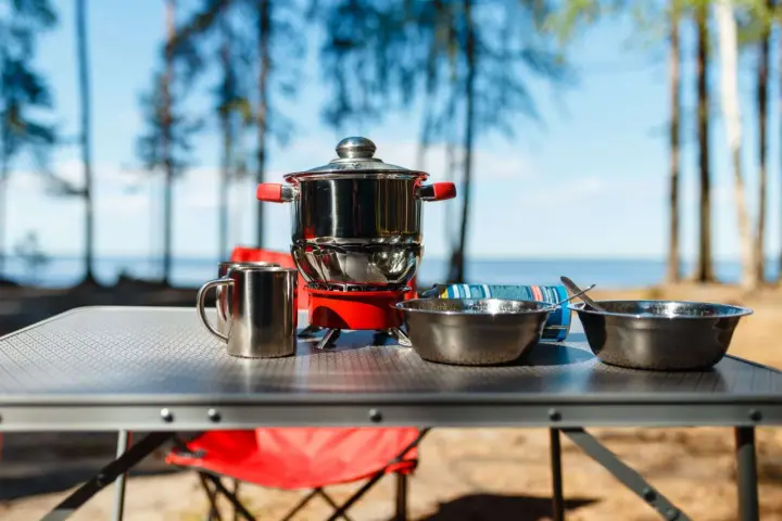 A complete camping kitchen setup with a stove, pots, bowls, and mugs on a table, ready for outdoor cooking by a lake.