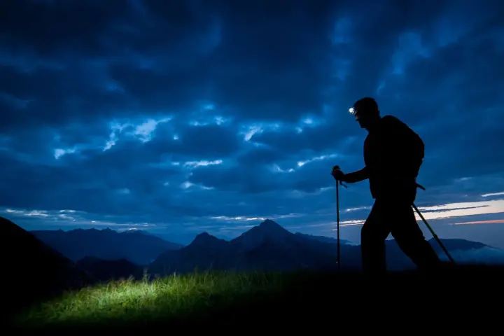 Silhouette of a hiker with a headlamp on, trekking poles in hand, on a night hike through a dramatic, cloudy sky over mountains.