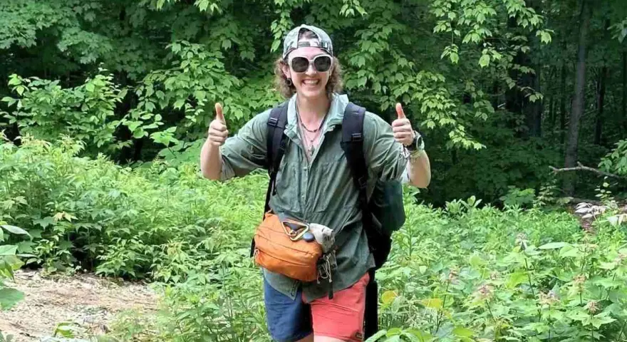 A hiker in a green, breathable hiking shirt material, gives a double thumbs-up in a lush forest.