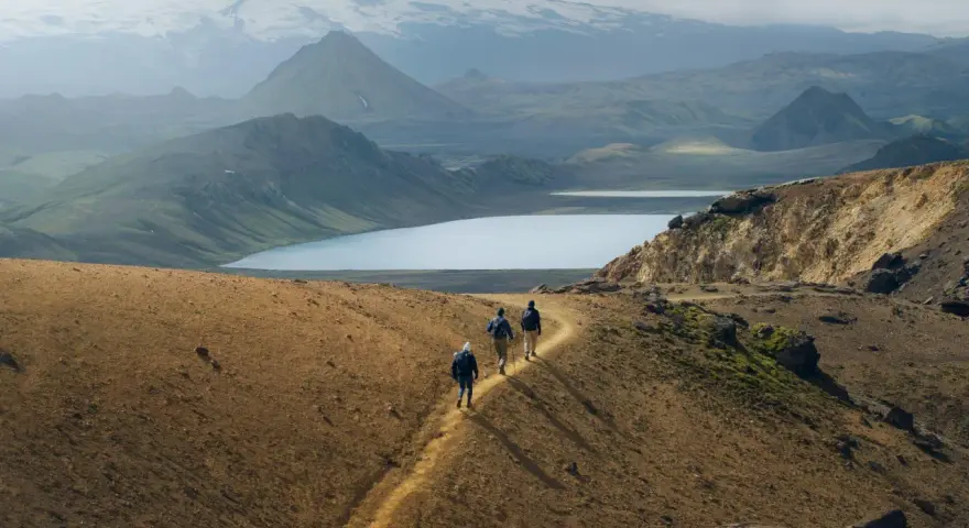 Hikers traverse a golden path on a mountain ridge, with a vast, serene lake and snow-capped peaks in the distance. This iconic landscape hints at the epic scale of how long is the Laugavegur trail.