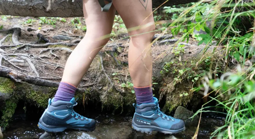 A hiker steps through a stream, testing how to waterproof hiking boots. Their blue boots and purple socks are ready for any wet trail.