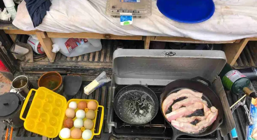 A camp kitchen checklist in action: bacon sizzles in a cast-iron pan, eggs are ready in a yellow carrier, and a portable stove is set up.