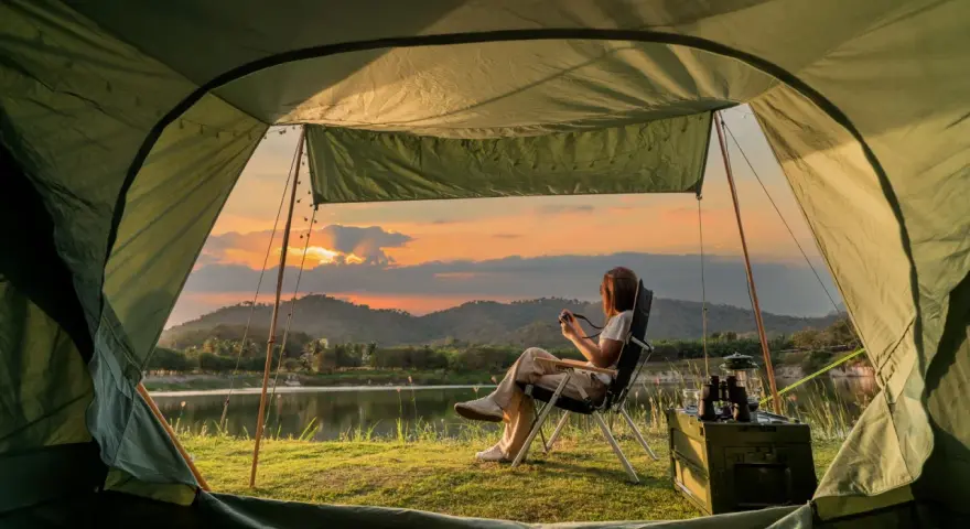 A woman enjoys solo camping by a lake at sunset, holding a camera. The tent offers a cozy view of the serene landscape.