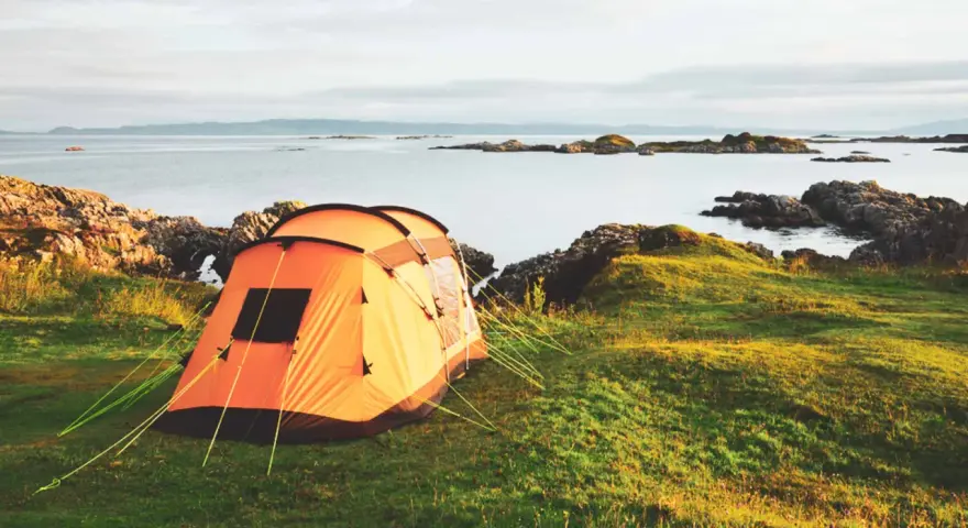 An orange tent is set up on a grassy hill overlooking a calm sea with rocky islands. This is a perfect spot for tent camp sites.