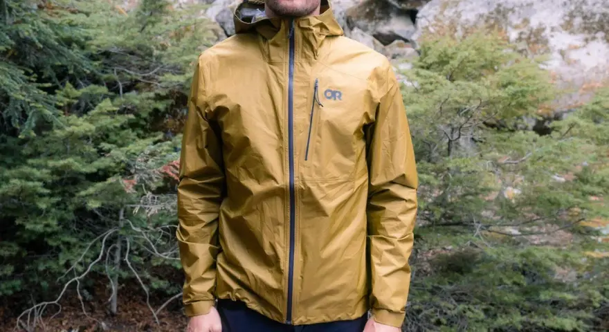 Man wearing a golden ultralight waterproof jacket with an OR logo, standing in a forest.