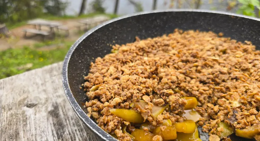 A warm, bubbling campfire apple crisp in a pan, ready to be enjoyed outdoors by a lake.