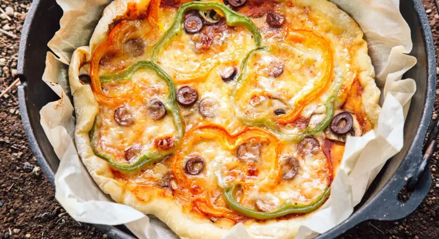 A delicious dutch oven pizza camping treat, topped with melted cheese, colorful bell peppers, and olives, baked to perfection on parchment paper.