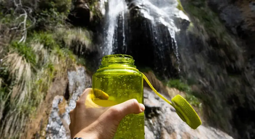 A hand holds a bright green water bottle, ready for a refreshing drink after a long hike. Behind it, a beautiful waterfall cascades down a rocky cliff, surrounded by lush greenery.