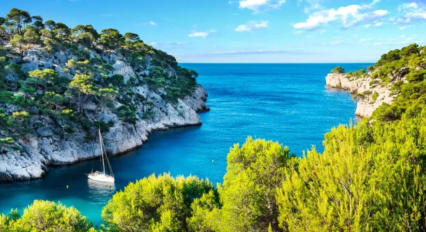A sailboat anchors in a serene cove, surrounded by lush green trees and dramatic cliffs. This idyllic scene evokes the feeling of a perfect camping trip in Cassis.