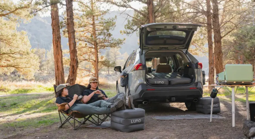 Two people enjoy a meal in camping chairs next to a car with an open trunk, showcasing clever car camping hacks with an inflatable mattress and a portable stove.